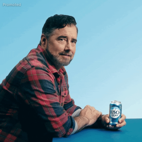 Sponsored gif. We zoom in on Gerald Downey seated in side profile in a red and black plaid shirt, holding a can of Busch Light beer on the table in front of him. He looks at us with a furrowed brow and confused expression as a single question mark appears next to him.