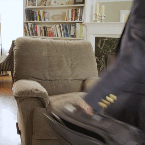 Video gif. A person in a business suit plops belly-first onto a recliner chair, then looks back at us and crosses legs up as text appears, "I'm exhausted."