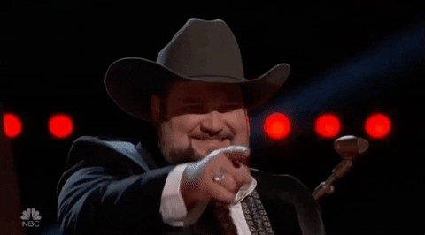 Reality TV gif. A contestant on The Voice is wearing a cowboy hat and he looks very pleased as he points at someone and grins.