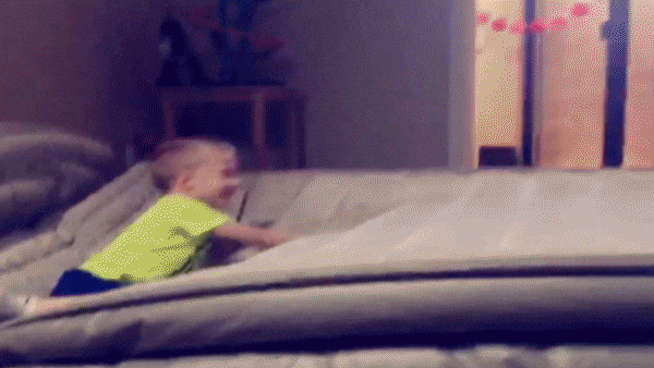 Video gif. Kid is crawling on an air mattress and laughing. A person jumps on the other side and sends him flying through the air, making his whole body flip comedically and land on the ground.