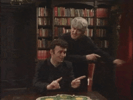 TV gif. In a room with a large bookshelf, Dermot Morgan as Ted from Father Ted stands behind a seated young man. Ted rocks back and forth with frustrated energy, pointing from side to side in rhythm as if singing a song, and the young man does the same without quite as much enthusiasm.