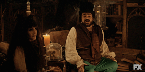 TV gif. Matt Berry as Laszlo and Natasia Demetriou as Nadja sit next to each other on separate arm chairs as Laszlo leans forward and points in agreement with someone off screen.