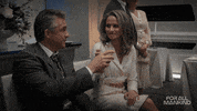 TV gif. Seated on an indoor bench and dressed in formal attire, a man and woman on For All Mankind smile at each other and clink their glasses, then take sips.