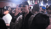 Video gif. Man stands in a crowd and wears a swaggy fit with black shades, a black cap, and an army green jacket on top of a black zip up jacket. He holds up his hand in front of his face and spreads his fingers to casually gesture, "What's up?"