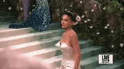 Met Gala 2024 gif. Slow motion close up of the top of Kylie Jenner's Oscar de la Renta champagne-colored strapless column gown featuring a pointed bust. She also wears a pale pink rose in a smooth mid bun.