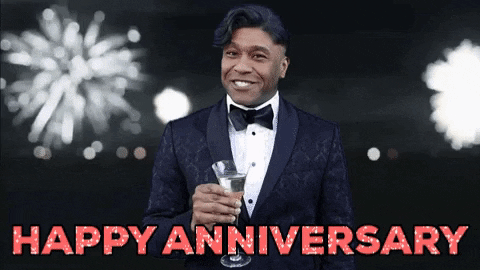 Meme gif. A man holds a glass full of champagne, motion to us with a "cheers" motion, in front of a background of bursting fireworks, in the style of the Leo DiCaprio "Great Gatsby" meme. The words "Happy Anniversary" appear as text.