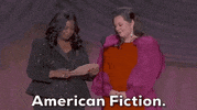 Oscars 2024 GIF. Melissa McCarthy and Octavia Spencer stand in front of the microphone. Spencer opens the envelope and announces the winner for Best Adapted Screenplay. She reads the envelope and looks up, saying, "American Fiction" and McCarthy reacts by sharply inhaling with excitement.
