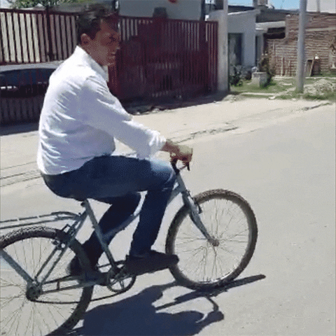 Video gif. Argentina's Economy Minister Sergio Massa rides a bike on an empty street, smiling and giving a thumbs up, then standing up on the pedals. 