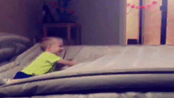 Video gif. Kid is crawling on an air mattress and laughing. A person jumps on the other side and sends him flying through the air, making his whole body flip comedically and land on the ground.