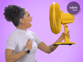 Video gif. Woman holds a big yellow fan to her face and fanning herself while saying, "Que Calor!"