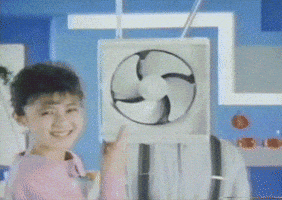 TV gif. As a woman points at an electric fan suspended from the ceiling, we zoom in to the fan's right. A man in suspenders leans out from behind the fan and mischievously covers his mouth.