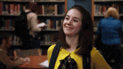 TV gif. Alison Brie as Annie Edison. She grins dorkily, leaning back on a table and begins to wave at someone but realizes the person isn't waving at her and she awkwardly touches her hair instead.