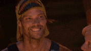 Reality TV gif. David Genat grins wide on Australian Survivor while smoke drifts in the background.