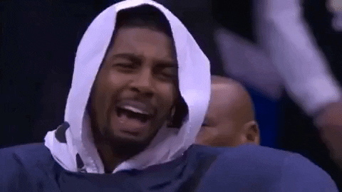 Sports gif. Kyrie Irving of the Cleveland Cavaliers sits on the sidelines with his hood up. He laughs hard and lowers his head to hide his laughter.