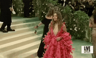 Met Gala 2024 gif. We zoom in on Jessica Biel waving and smiling at camera, wearing a hot pink Tamara Ralph Couture full-length gown with a deep-v neckline. The full billowing skirt flares out to a dramatic train. She wears a billowing shrug that matches the pattern of her dress around her back and arms.  