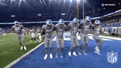 Sports gif. Five Detroit Lions football players in uniform do a can-can line on the end zone.