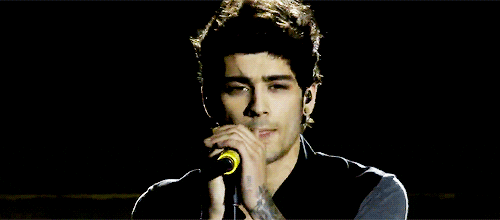 Celebrity gif. Zayn Malik licks his lips and pushes his microphone off to the side, then looks down.