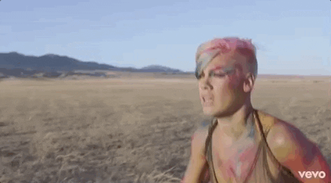 Music video gif. From the video for Try by Pink, the singer Pink runs in a flat grassy field, wearing a green bra, skin dappled with colors.