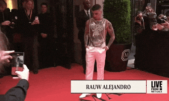 Met Gala 2024 gif. Rauw Alejandro wearing a golden sleeveless top and white satin pants, an ensemble by Ludovic de Saint Sernin, turns slowly, adjusting nervously, fully surrounded by cameras.
