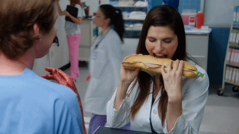 TV gif. Lake Bell as Dr. Cat Black in Children's hospital stands in front of a doctor with bloody hands. He holds a long submarine sandwich and rips a bite out of the side of it while the other doctor talks to her.