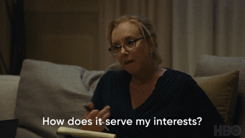 TV gif. J. Smith-Cameron as Gerri on Succession sits on a couch with a notepad and asks, with her head tilted sideways, "How does it serve my interests?" which appears as text.