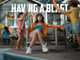 Sponsored gif. Aubrey Plaza is at a pool party sitting in a patio chair with her legs crossed while holding a Mountain Dew Baja Blast in her right hand. Two kids on either side are smacking her on top of her head with pool noodles ruffling her hair. She couldn't care less as she looks down at us, raises her left hand nonchalantly and says, "Having a blast."