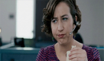 Movie gif. Kristen Schaal as Susana from Dinner for Schmucks has her work headphones on and is eating half a sandwich while she rolls her eyes and smacks her lips in disgust before taking another bite.