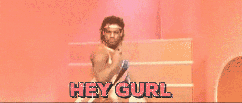 Video gif. We zoom in on a man wearing a gold lamè headband and spandex tank top as he looks over his shoulder at us with an intense gaze. Text, "Hey gurl."