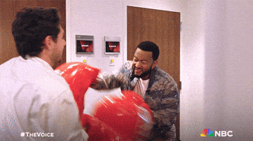 Celebrity gif. John Legend and Niall Horan are wearing giant inflatable boxing gloves and they're fighting each other.