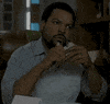 Celebrity gif. Ice Cube sits looking skeptical, chewing slowly on the sandwich that he is holding.