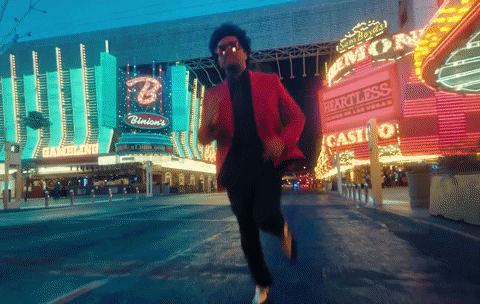 Celebrity gif. Singer The Weeknd (pronounced Weekend) runs wildly down the Las Vegas strip wearing a pink suit jacket and sunglasses. His arms flail wildly around him as he runs.