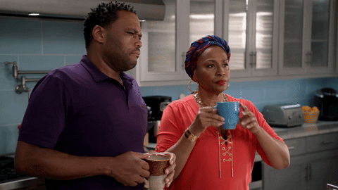 TV gif. Anthony Anderson as Dre Johnson and Jenifer Lewis as Ruby Johnson on Black-ish stand in the kitchen staring at someone with judging expressions. They lift their coffee mugs to their mouths and take a sip of coffee at the same time as they continue to stare.