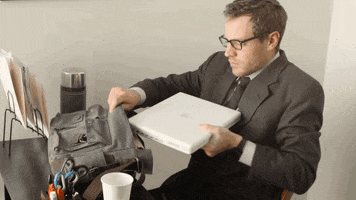 Video gif. Man wearing a suit and sitting at a desk struggles to stuff a MacBook into a messenger bag, from the music video for "Worth" by Clique.