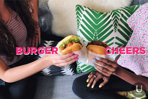 Video gif. Two women sit next to each other on a sofa and move their burgers towards each other like they’re clinking glasses together. Text, “Burger cheers.”