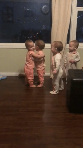 Video gif. Four babies all stand up. Two babies in pink hug each other while the other two awkwardly stand and wait their turn. One of the babies in white taps a baby in pink and they hug. The four babies then go around, hugging each other and smiling with excitement as they pat each other’s backs. 