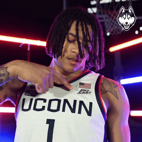 Sports gif. Solomon Ball, a UConn Huskies basketball player, points to a logo on his jersey that reads "Big East," looking at the logo then back at us with a confident expression. 