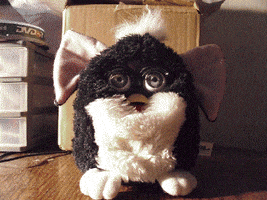 Stop motion gif. Black and white Furby drops its ears, then its outer furry layer, then scoots its robotic body toward us. The image becomes ominously red-tinted and text appears, "yuuuuum."