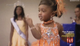Reality TV gif. Toddler wearing pageant gear smiles and spins in a circle while flipping us off.