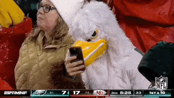 Sports gif. A person in an Eagles mascot costume holds a cell phone in front of their face and cocks its head to look at the screen as rain pours down on all the fans in the stands around it. 