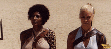 Movie gif. Pam Grier as Mamawi and Margaret Markov as Boudicca in The Arena raise weapons to a cheering crowd.