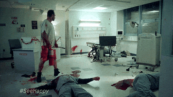 TV gif. Wearing a bloody hospital gown and holding a fire extinguisher, Christopher Meloni as Nick Sax in Happy! looks around at the gory scene around him and says, “I can explain.”