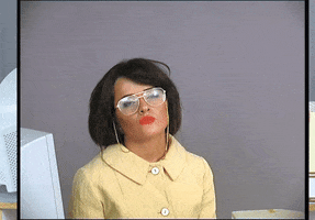 Video gif. A woman sits in a mundane, stereotypical office cubicle. She’s dressed in an old fashioned yellow coat and wears big aviator glasses that have a chain attached to them. She sits there boredly, staring into space. She then looks at us with a deadpan expression. 