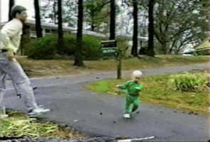 Video gif. Baby is in a driveway and they fall, catching themselves on their hands. They can't find their center of balance and they do a handstand and flip over, an adult running to help.