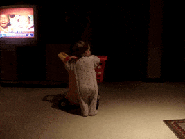 Video gif. A baby plays with a toy shopping cart as they wobble over and the cart topples on top of them. 