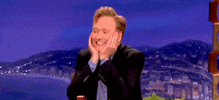 TV gif. Conan O'Brien cups his face with his hands, smiling and dancing excitedly in his seat.
