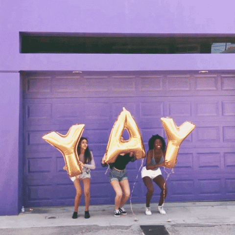 Video gif. Three women jump up in front of a purple garage door, holding gold balloons that spell out, "Yay."