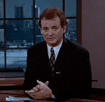 Movie gif. Bill Murray as Phil from Groundhog Day speaks to us from a news desk, and begins to smile as he crosses his fingers. Text, "Fingers crossed."