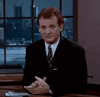 Movie gif. Bill Murray as Phil from Groundhog Day speaks to us from a news desk, and begins to smile as he crosses his fingers. Text, "Fingers crossed."