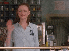 TV gif. Alexis Bledel as Rory in Gilmore Girls waves toward us with a taut smile.