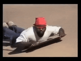 Meme gif. From the Dank Ass Sandboarding Son meme, a man in sunglasses and a red bandanna bodysurfs across a sand dune on a plank, and gives a hang loose gesture with a chill expression as he passes us.